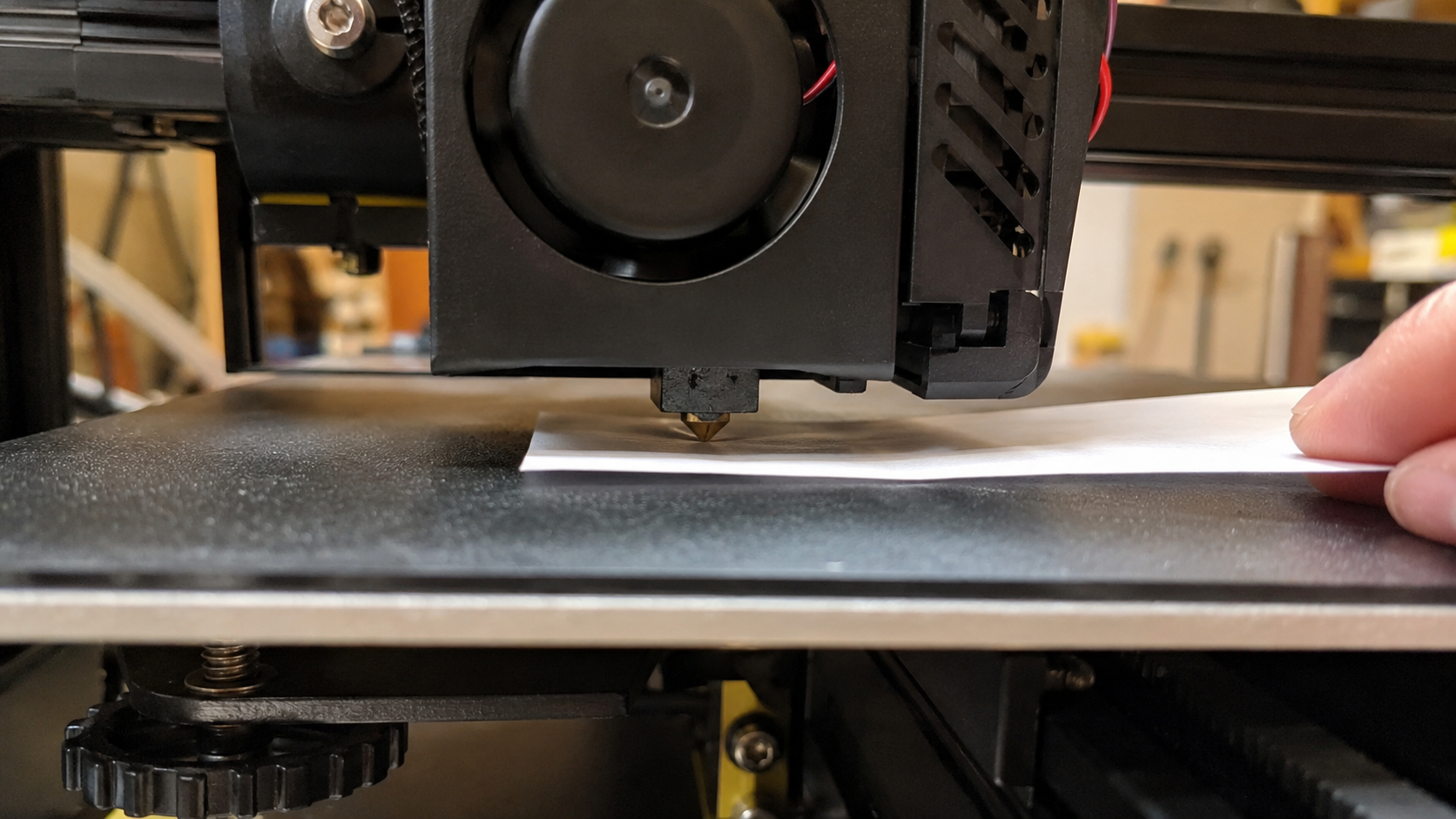 Side view of a printer nozzle with a sheet of paper between it and the build plate during manual bed leveling, showing the correct paper drag technique for 3D printer bed leveling